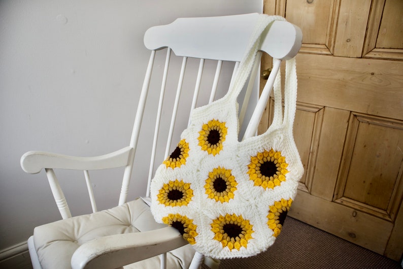 Puede incluir: Una bolsa de mano de crochet blanca con un patr&oacute;n de girasol. La bolsa est&aacute; colgada de una mecedora blanca.