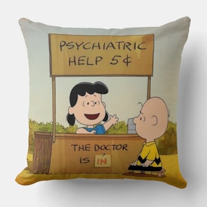 Puede incluir: Una almohada de color amarillo y marrón con una imagen de dibujos animados de Peanuts. La imagen muestra a Lucy sosteniendo un cartel que dice "Psychiatric Help 5$" y a Charlie Brown sentado frente al cartel. El cartel también dice "The Doctor is in".