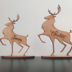 Może przedstawiać: Dwie drewniane figurki renifer&oacute;w z wygrawerowanymi imionami. Jedna z nich to "James", a druga "Charlotte". Renifery są brązowe i stoją na okrągłych podstawach, idealne do dekoracji świątecznych.