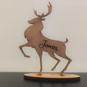 Może przedstawiać: Brązowa drewniana figurka jelenia z wygrawerowanym imieniem "James" z boku. Jeleń stoi na małej, owalnej drewnianej podstawie. Jeleń jest ustawiony z jedną uniesioną nogą, co nadaje mu dynamiczny wygląd.