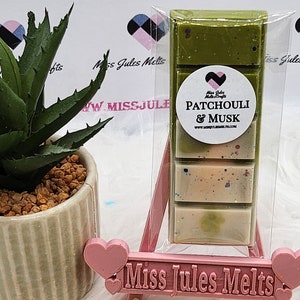 Puede incluir: Una barra de cera fundida en un envase de plástico transparente con un diseño verde, blanco y rosa. La barra está etiquetada como "Patchouli & Musk" y está empaquetada por "Miss Jules Melts & Crafts".