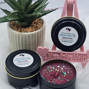 Puede incluir: Dos latas de velas negras con tapas doradas. La vela de la izquierda está etiquetada como "Fresh Mint & Rhubarb Crackle Candle". La vela de la derecha está llena de cera de vela roja con purpurina dorada. Las velas están sobre una superficie blanca y esponjosa.