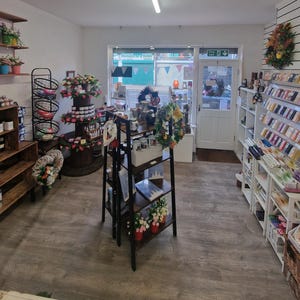 May include: A retail shop interior with wooden shelves displaying various products, including floral arrangements, mugs, and decorative items. The shop has a welcoming atmosphere with a variety of merchandise and a door leading outside.