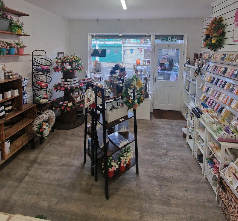 May include: A retail shop interior with wooden shelves displaying various products, including floral arrangements, mugs, and decorative items. The shop has a welcoming atmosphere with a variety of merchandise and a door leading outside.