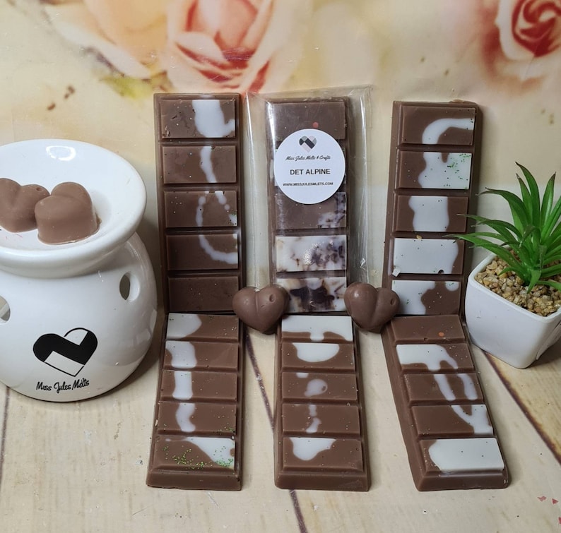 May include: An assortment of scented wax melts in various shapes and designs. The melts are primarily brown with white and green accents. The image also includes a white ceramic oil warmer and a small potted plant.