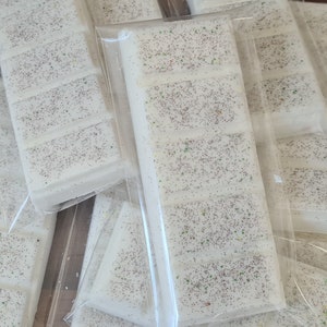 May include: Close-up of several white wax melt bars, each divided into sections and sprinkled with silver and colourful glitter. The bars are packaged in clear plastic, suggesting a home fragrance product.