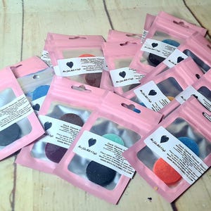 Könnte beinhalten: Eine Sammlung von rosa, wiederverschließbaren Beuteln, die jeweils ein farbenfrohes, rundes Produkt enthalten. Die Produkte sind in verschiedenen Farben erhältlich, darunter Blau, Lila und Orange. Jeder Beutel hat ein weißes Etikett mit Text und einem schwarzen Herzlogo.