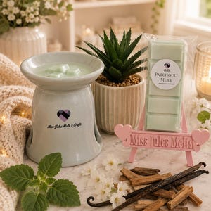 Pode incluir: Um aquecedor de cera de cerâmica verde-claro com o logótipo Miss Jules Melts & Crafts, uma barra de cera a condizer com a etiqueta Patchouli Musk numa embalagem transparente sobre um suporte rosa em forma de coração, folhas verdes, vagens de baunilha e pequenas flores brancas.