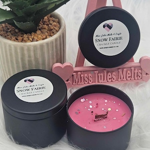 May include: Two black metal candle jars with pink lids. The jars are labelled "Miss Jules Melts & Crafts Snow Fairie Crackle Candle" and have a pink heart design. One jar is filled with a pink candle with glitter and a chocolate piece on top.