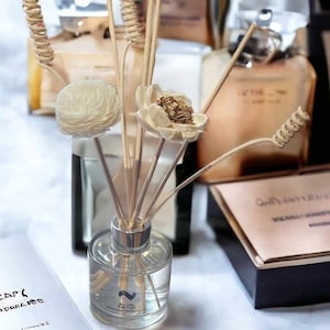 May include: A clear glass diffuser bottle with a silver cap and dried flowers. The diffuser is filled with a clear liquid and has reeds sticking out of the top. The bottle is sitting on a white surface.
