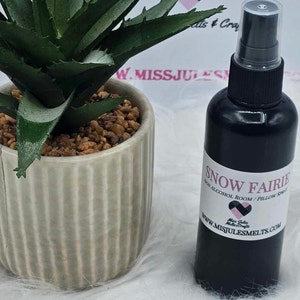 May include: A black bottle with a white label that reads "SNOW FAIRIE NON ALCOHOL ROOM/PILLOW SPRAY" and a website address. The bottle is sitting on a white surface next to a potted succulent.