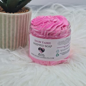 Puede incluir: Un tarro de jabón batido rosa Snow Fairie con una apariencia batida texturizada. El tarro tiene una etiqueta con el texto "Snow Fairie Whipped Soap" y la dirección del sitio web www.MISSJULESMELTS.COM. El tarro está sobre una superficie blanca y esponjosa.