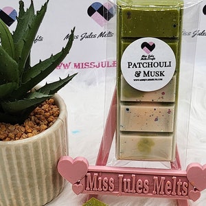Puede incluir: Un paquete de ceras perfumadas con el texto "Patchouli & Musk" sobre un caballete rosa. Las ceras están divididas en secciones de colores verde y beige. A la izquierda hay una pequeña planta suculenta en maceta.