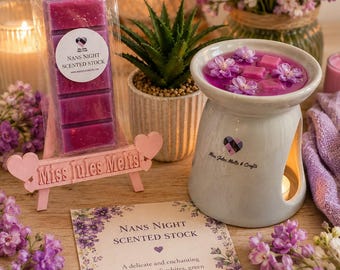 Op de afbeelding: Een geurwaxbar met het label Nans Night Scented Stock in een plastic verpakking, een witte keramische waxbrander met een hartlogo en een kaart met de tekst Nans Night Scented Stock. De brander bevat gesmolten paarse wax en bloemvormige stukjes.