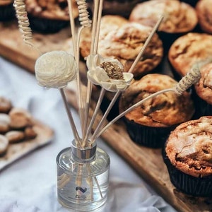 Może przedstawiać: Szklana butelka dyfuzora z przezroczystego szkła z drewnianymi patyczkami i białymi akcentami kwiatowymi. Dyfuzor stoi na drewnianej powierzchni z różnymi muffinkami w tle.