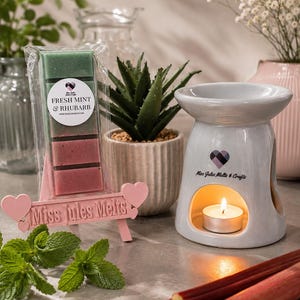 May include: A wax melt bar labeled Fresh Mint & Rhubarb in a clear wrapper, displayed on a pink heart-themed stand, alongside a white ceramic oil burner with a lit tea light candle, featuring the text Miss Jules Melts & Crafts on both items.