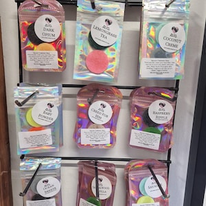 Puede incluir: Exhibición de discos ambientadores para aspiradora con varios aromas, incluyendo "Dark Opium", "Lemongrass Tea" y "Coconut Creme". Cada disco está empaquetado en una bolsa transparente iridiscente y se exhibe en un estante de metal negro.