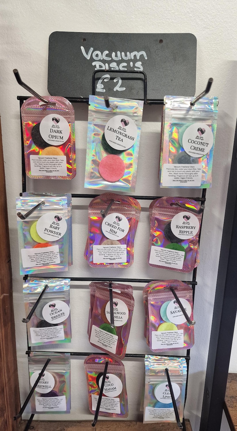 May include: A display of vacuum freshener discs in various scents, including "Dark Opium," "Lemongrass Tea," and "Coconut Creme." Each disc is packaged in a clear, iridescent pouch and displayed on a black metal rack. The sign above reads "Vacuum Disc's."