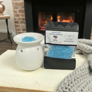 Puede incluir: Un quemador de aceite de cerámica blanca con cristales azules y una bolsa negra de "Clean Linen Sizzling Crystals". La imagen está ambientada frente a una chimenea y una manta gris tejida.