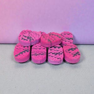 May include: Eight pink bath bombs with a dark grey marbled pattern. The bath bombs are stacked against a light purple backdrop. Each bath bomb is approximately 5 cm in length.