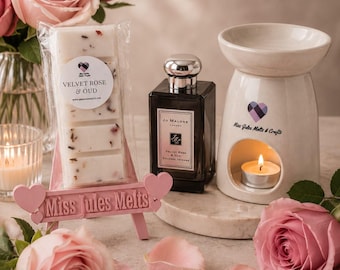 May include: A wax melt bar labeled Velvet Rose & Oud, a dark glass perfume bottle, and a white ceramic oil burner with a lit tea light candle. A pink easel displays the wax melt, surrounded by pink roses, petals, and a glass vase of flowers.