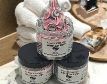 Może przedstawiać: Trzy przezroczyste słoiki mydła w piance Black Cherry z czarnymi wieczkami. Mydło jest wirujące z różowymi i szarymi kolorami. Każdy słoik ma etykietę z nazwą produktu i adresem strony internetowej. W tle stos białych ręczników.