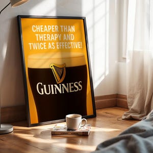 Może przedstawiać: Oprawiony plakat Guinnessa z kuflem stoutu, tekstem "Cheaper Than Therapy And Twice As Effective!" i logo Guinness. Filiżanka kawy i książka leżą na podłodze przed plakatem.
