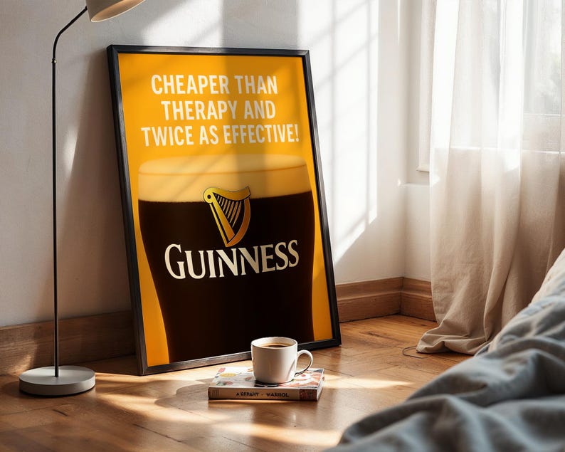 Może przedstawiać: Oprawiony plakat Guinnessa z kuflem stoutu, tekstem "Cheaper Than Therapy And Twice As Effective!" i logo Guinness. Filiżanka kawy i książka leżą na podłodze przed plakatem.