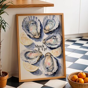 Op de afbeelding: Een ingelijst aquarel schilderij van vijf oesters en citroenschijfjes op een witte achtergrond. De tekst "Shuck It Up!" is geschreven in zwarte letters.