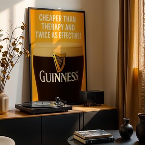 Może przedstawiać: Oprawiony plakat Guinness z napisem &bdquo;Cheaper Than Therapy And Twice As Effective&rdquo; jest wyświetlany nad gramofonem. Plakat przedstawia kufel Guinnessa. Czarny gramofon stoi na drewnianej szafce.