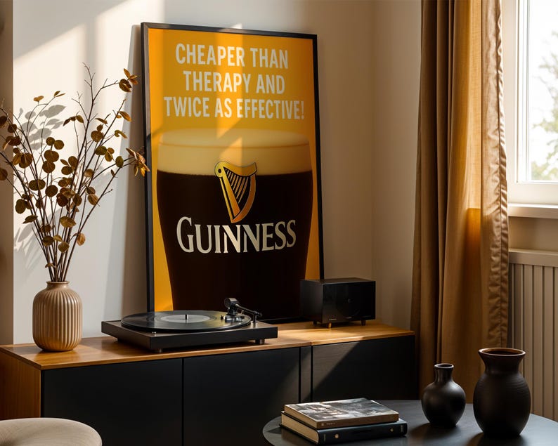 Może przedstawiać: Oprawiony plakat Guinness z napisem &bdquo;Cheaper Than Therapy And Twice As Effective&rdquo; jest wyświetlany nad gramofonem. Plakat przedstawia kufel Guinnessa. Czarny gramofon stoi na drewnianej szafce.