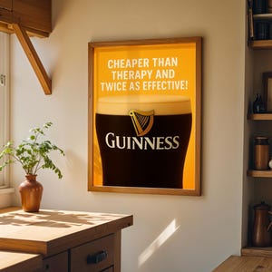 Może przedstawiać: Oprawiona reklama Guinnessa z napisem "Tańsze niż terapia i dwa razy skuteczniejsze!" Obraz przedstawia kufel Guinnessa z logo marki. Rama jest w kolorze drewna, a tło to kuchnia.