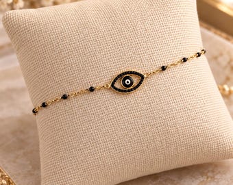 Puede incluir: Una delicada pulsera de cadena dorada con un dije central en forma de ojo con esmalte negro y una pupila blanca. La cadena está adornada con pequeñas cuentas negras espaciadas uniformemente, creando una pieza de joyería refinada.