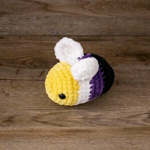 May include: A crocheted bee plush toy in yellow, white, purple, and black. The bee has white wings, a yellow body, and a black eye. A soft, cuddly toy.