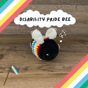 Puede incluir: Una abeja de peluche hecha a mano con cuerpo negro, rayas arcoíris y alas blancas. El texto "DISABILITY PRIDE BEE" está en una nube blanca. Una pequeña bandera con rayas arcoíris está en la esquina inferior izquierda.