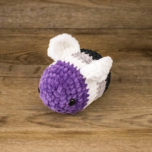 May include: A handmade crochet bee in purple, white, gray, and black. The plush toy has black safety eyes and is made of soft, textured yarn. The bee is lying on a wooden surface.