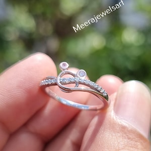 May include: A silver ring with a unique design, featuring small clear stones and a central knot-like detail. The ring is held in a hand, showcasing its delicate craftsmanship. The ring is a piece of jewelry.