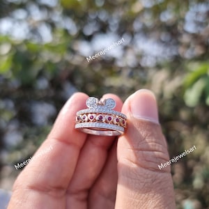 May include: A multi-band ring featuring a top section shaped like a mouse head with white stones, a middle band with a row of small red circular stones set within heart-shaped metal frames, and a bottom band encrusted with small white stones.