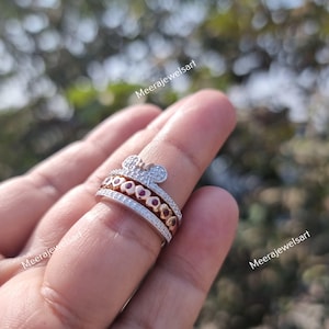 May include: A multi-band ring featuring a top band with a mouse-ear silhouette encrusted with clear stones, a middle band with a row of rose gold hearts containing red stones, and a bottom band paved with clear stones. The text Meerajewelsart is visible.