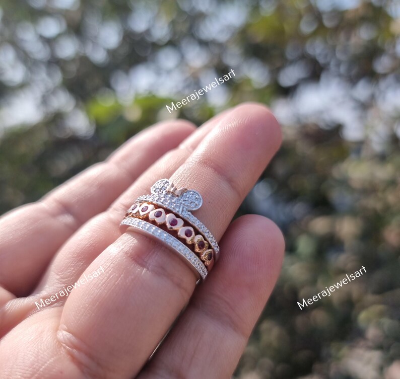 May include: A multi-band ring featuring a top band with a mouse-ear silhouette encrusted with clear stones, a middle band with a row of rose gold hearts containing red stones, and a bottom band paved with clear stones. The text Meerajewelsart is visible.