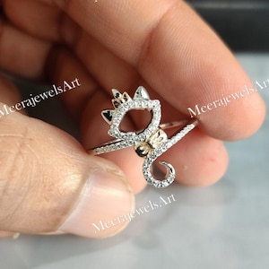May include: A silver ring in the shape of a cat, featuring a crown and a curled tail. The ring is adorned with small, clear gemstones. The cat's head and tail are outlined with the gemstones, and the crown is also detailed with them.