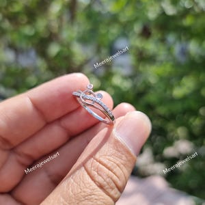 May include: A silver ring with a unique design, featuring small clear and pink gemstones. The ring has a double band and a decorative top with a heart-like shape. The ring is held in a hand against a blurred green background.