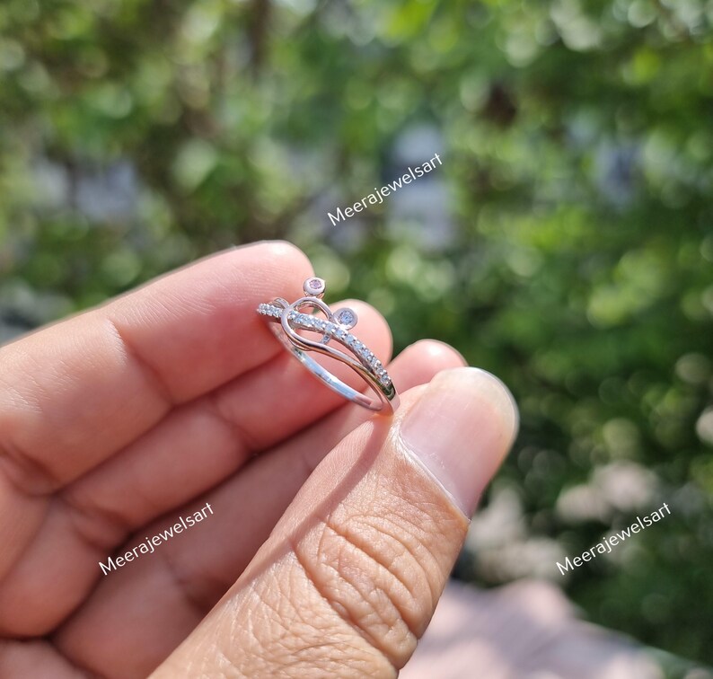 May include: A silver ring with a unique design, featuring small clear and pink gemstones. The ring has a double band and a decorative top with a heart-like shape. The ring is held in a hand against a blurred green background.