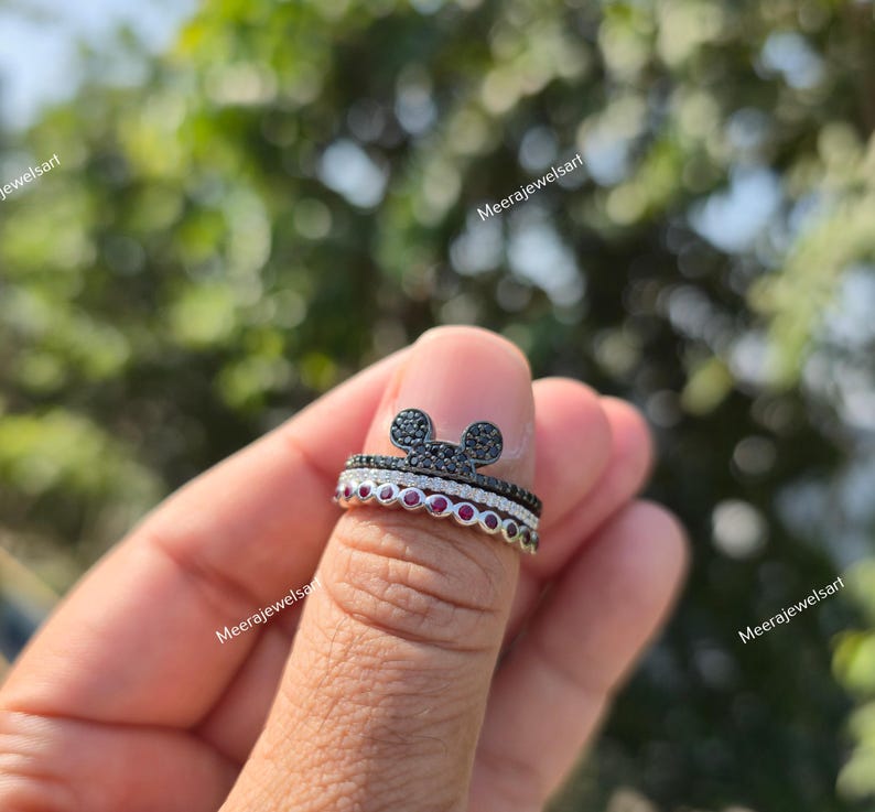 K&ouml;nnte beinhalten: Ein stapelbares Ring-Set bestehend aus drei Ringen. Der oberste Ring zeigt eine mit schwarzen Steinen besetzte Mausohr-Silhouette, der mittlere Ring ist eine d&uuml;nne Reihe klarer Steine und der untere Ring besteht aus einer Reihe runder roter Steine.