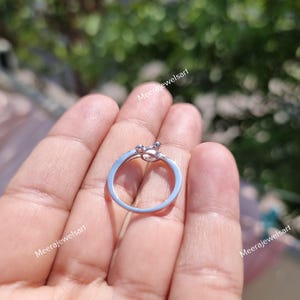May include: A silver ring with a unique design, featuring a small, decorative element at the top. The ring is held in a hand, showcasing its delicate details and craftsmanship. The ring's band is thin and appears to be made of a shiny metal.