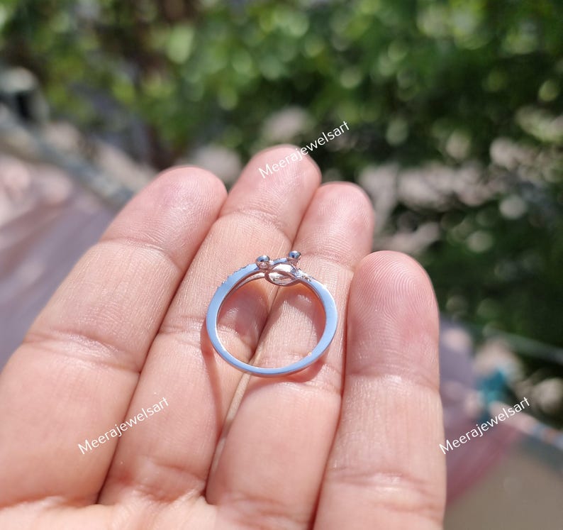 May include: A silver ring with a unique design, featuring a small, decorative element at the top. The ring is held in a hand, showcasing its delicate details and craftsmanship. The ring's band is thin and appears to be made of a shiny metal.