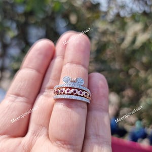 May include: A silver-toned ring featuring a top row of pav&eacute;-set clear stones, a middle row of small heart-shaped cutouts containing round red stones, and a bottom row of pav&eacute;-set clear stones, topped with a small bow-shaped accent piece.