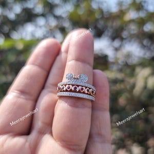 May include: A multi-band ring featuring a top band with a bow shape, a middle band with a series of small red heart-shaped stones, and two outer bands encrusted with tiny clear stones. The ring is crafted from a metallic material with a rose gold tone.
