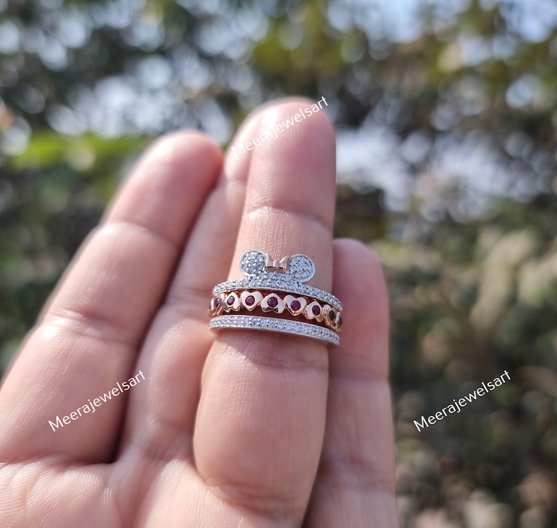 May include: A multi-band ring featuring a top band with a bow shape, a middle band with a series of small red heart-shaped stones, and two outer bands encrusted with tiny clear stones. The ring is crafted from a metallic material with a rose gold tone.