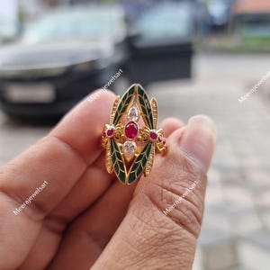 May include: A gold ring featuring a central oval red gemstone flanked by two pear-shaped clear stones, surrounded by green enamel leaf-shaped accents and small clear stones. The ring has a pointed, elongated design with intricate metal detailing.